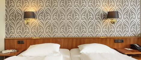Habitación de hotel con papel tapiz estampado, dos lámparas de noche, un cabecero de madera y una cama con ropa blanca.