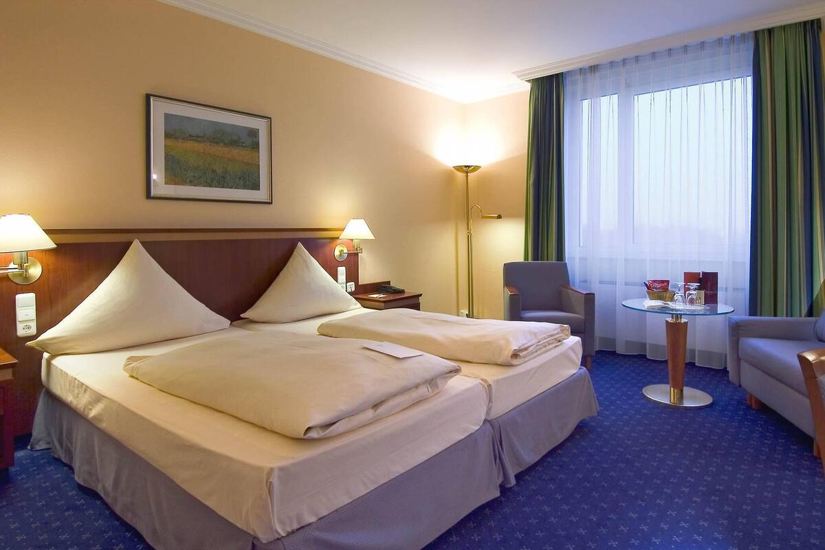 A hotel room with a double bed, two bedside lamps, a chair, a small table, and a window with curtains.