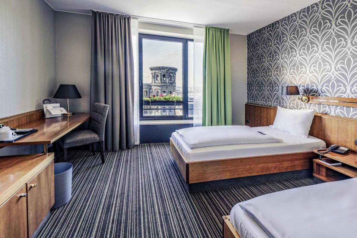 Habitación de hotel con cama, escritorio, silla, mesita pequeña, mesita de noche, lámpara, teléfono y vista a un edificio a través de una ventana.