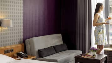 Habitación de hotel con cama, sofá gris, una mesita con un florero y ventana con cortinas.