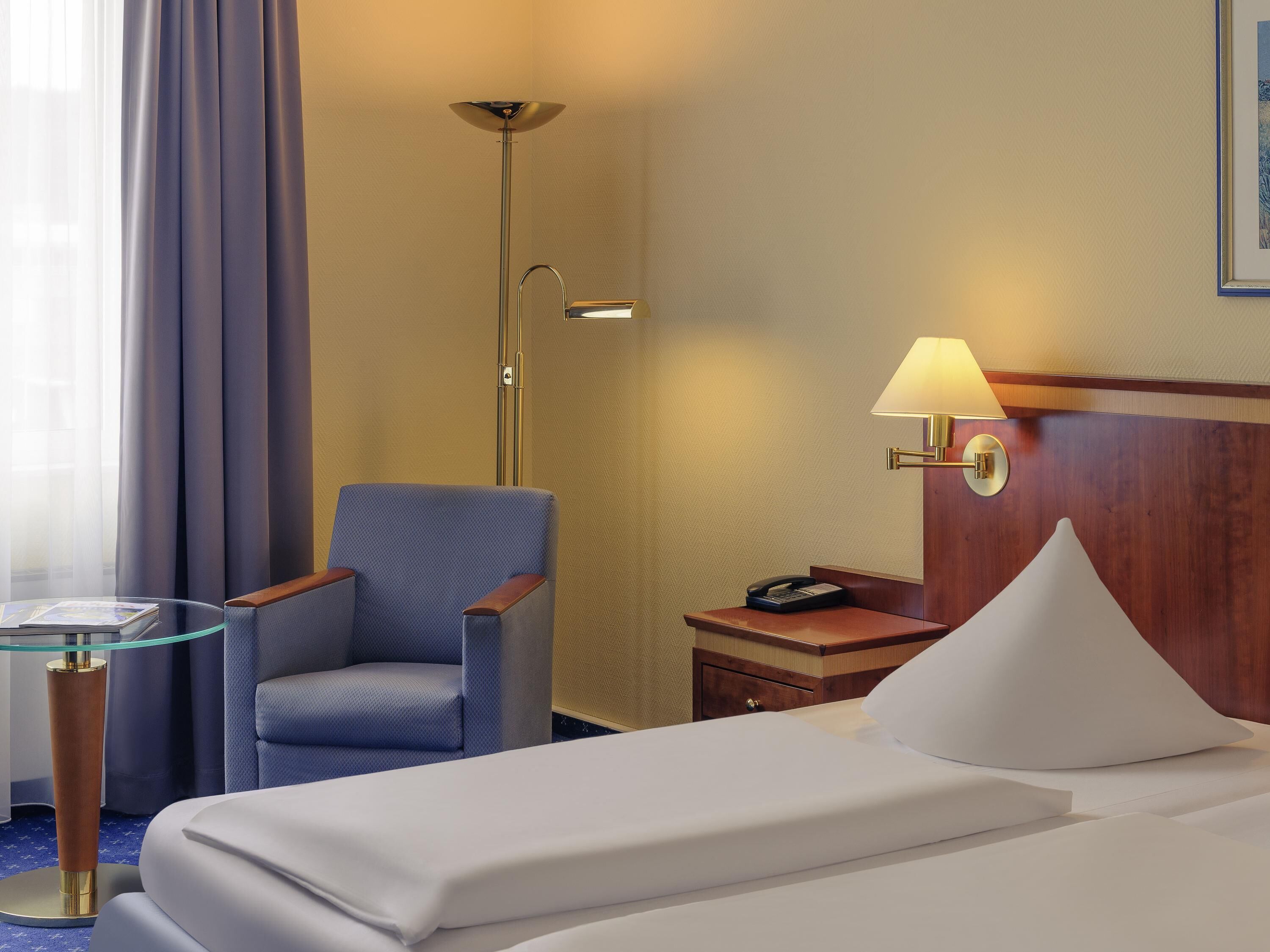 Ein Hotelzimmer mit einem Bett, einem Sessel, einem Nachttisch mit Lampe, einem Telefon und einem Fenster mit Vorhängen.