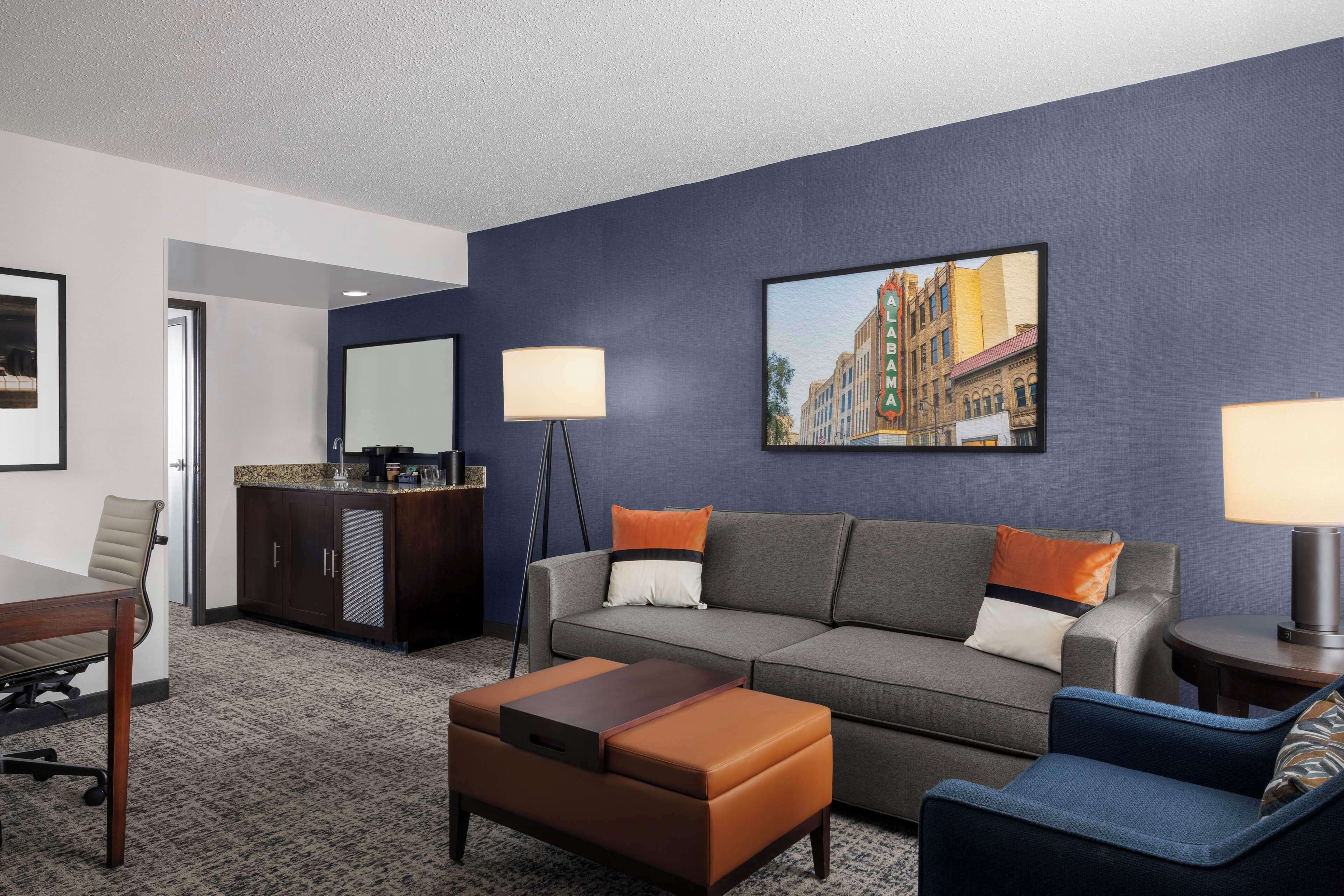 Una habitación de hotel moderna con un sofá gris, un sillón azul, una mesa de centro de madera, un mueble bajo de madera oscura y un cuadro con una vista urbana en la pared.