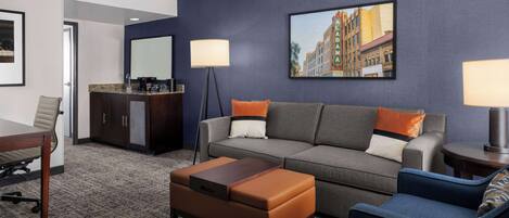 Una habitación de hotel moderna con un sofá gris, un sillón azul, una mesa de centro de madera, un mueble bajo de madera oscura y un cuadro con una vista urbana en la pared.