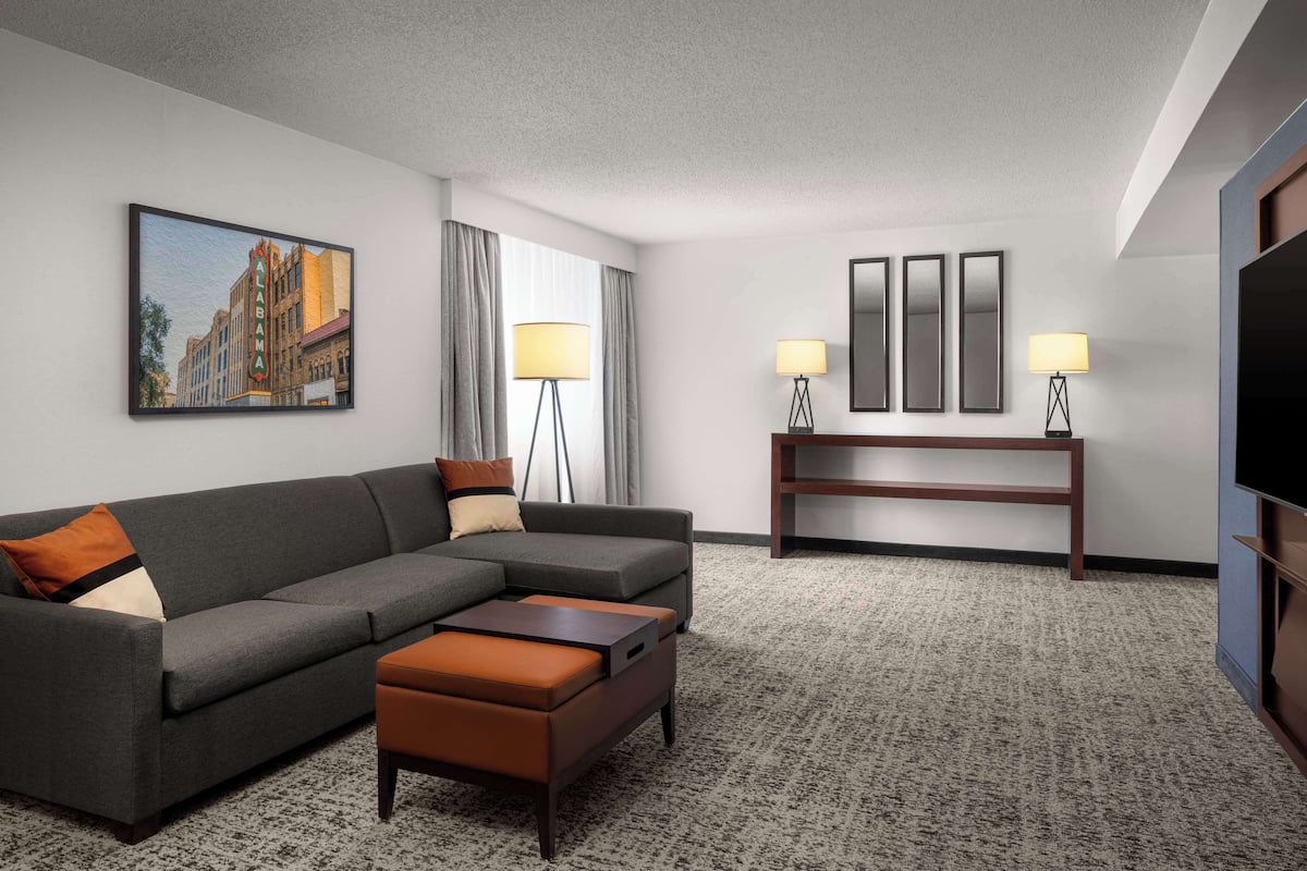 A modern hotel room with a grey sofa, a small ottoman, a wooden side table, and a flat-screen TV.