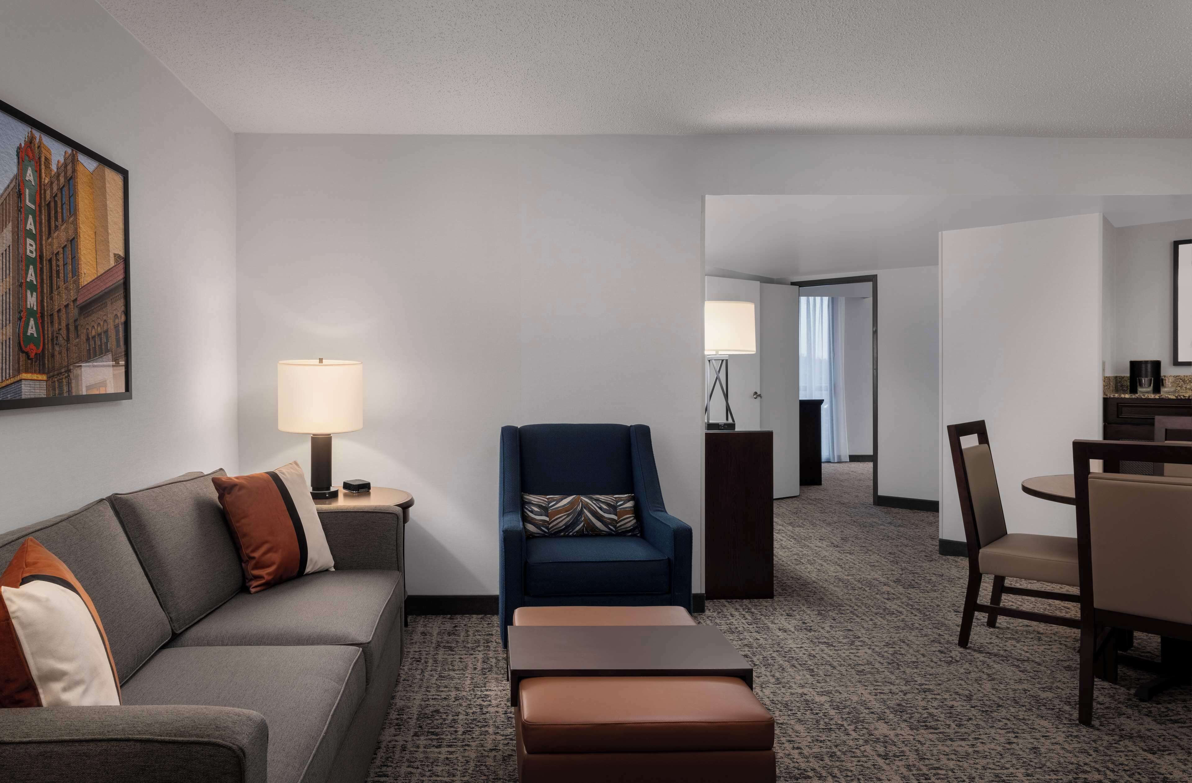 A hotel room with a gray sofa, a blue armchair, a small table, and a dining area with a chair and table.