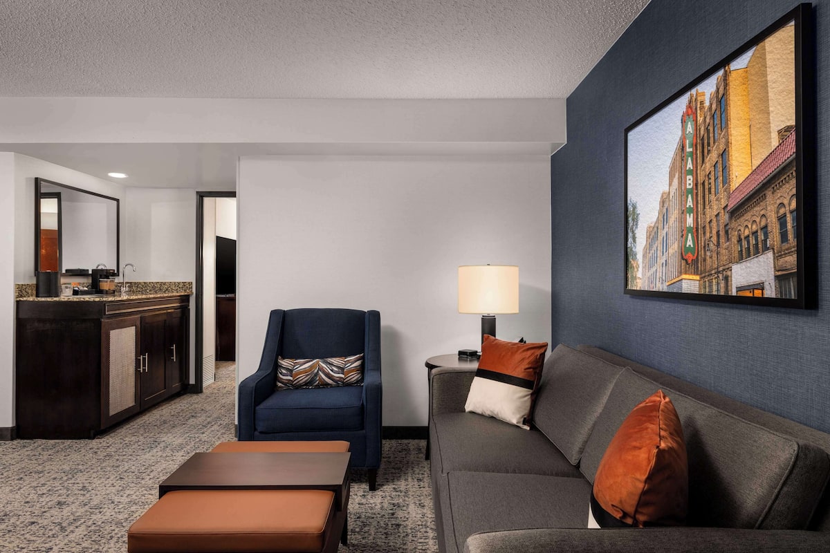 A hotel room with a living area featuring a sofa, armchair, and ottoman. A painting of a building is on the wall, and there is a small table with a lamp.