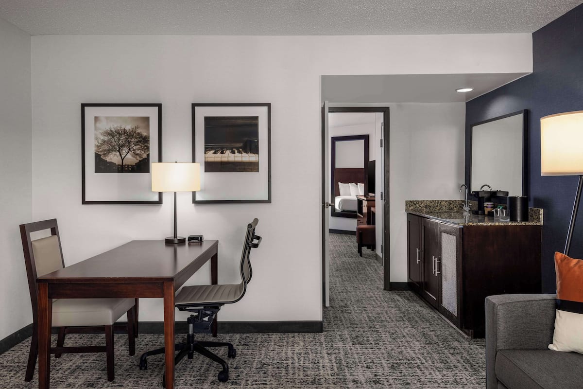 A hotel room with a dining table, two chairs, a lamp, framed pictures on the wall, a bed, a bathroom with a sink, and a sofa.