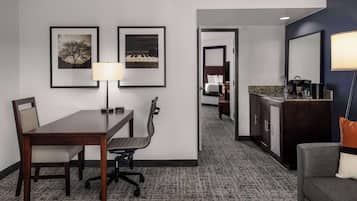 A hotel room with a dining table, two chairs, a lamp, framed pictures on the wall, a bed, a bathroom with a sink, and a sofa.
