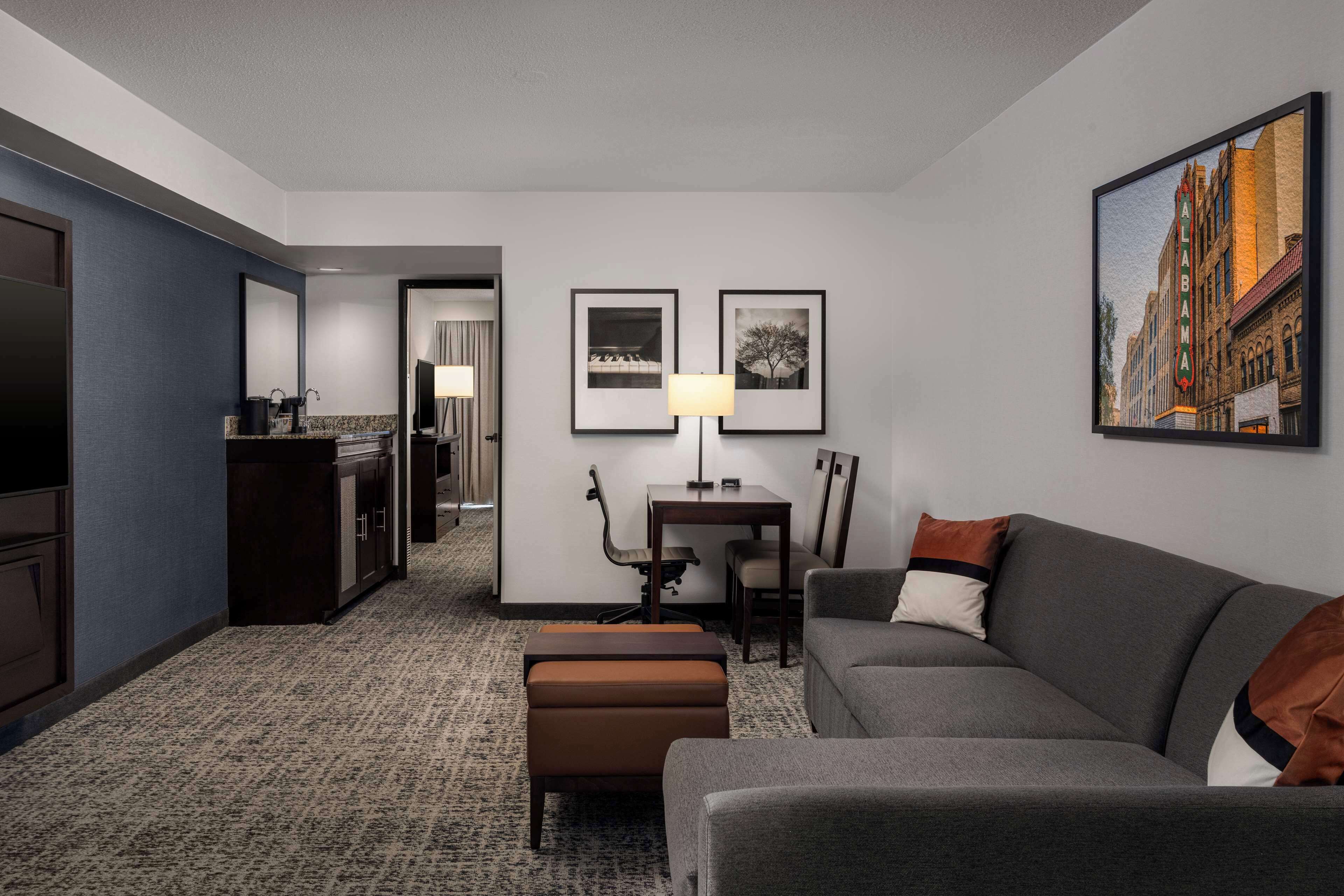 A hotel room with a grey sofa, a small table with a lamp, a framed picture of a building, and a carpeted floor.