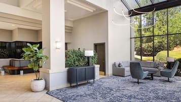 Un vestíbulo espacioso con un amplio ventanal, una zona de estar con sofá y sillones, una planta en maceta y un radiador.