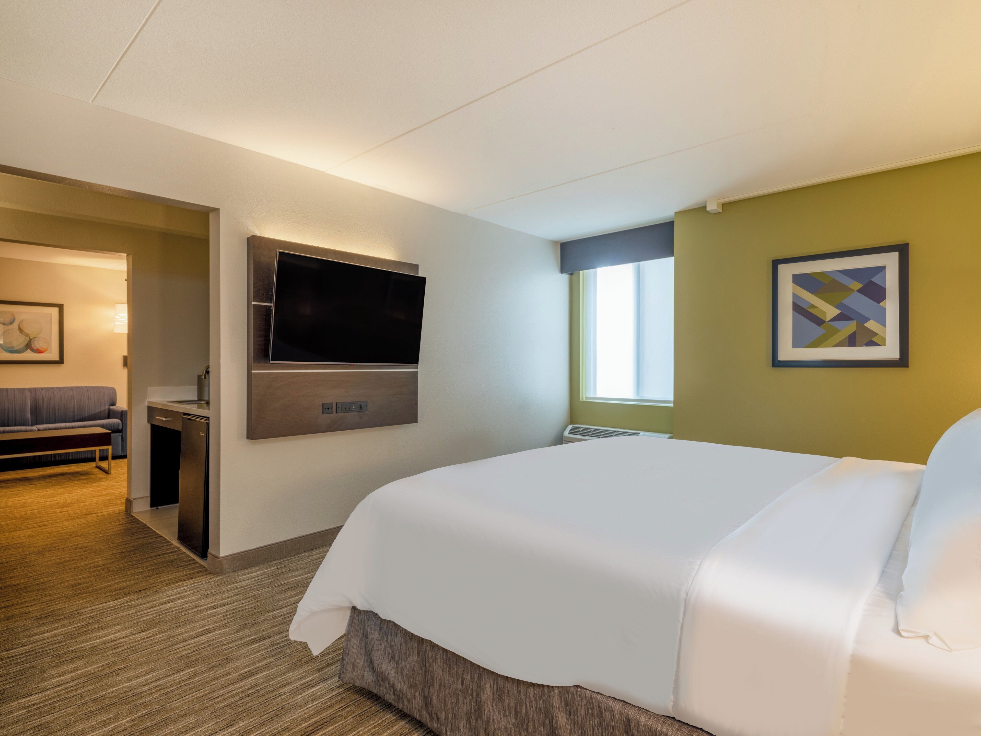 Una habitación de hotel con una cama grande, un televisor de pantalla plana en la pared, una kitchenette pequeña y una ventana con persianas.