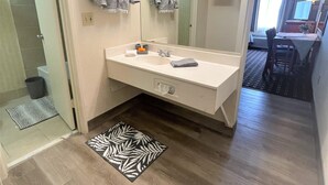 A hotel bathroom with a white sink, mirror, and vanity, a shower area, and a small rug. - FairBridge Extended Stay – a Kitchenette Hotel (Idaho Falls)