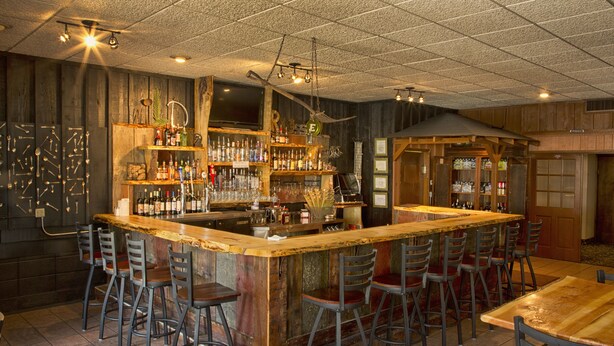 A bar area with a wooden bar counter, multiple stools, and a variety of bottles and glasses on shelves. - FairBridge Extended Stay – a Kitchenette Hotel (Idaho Falls)