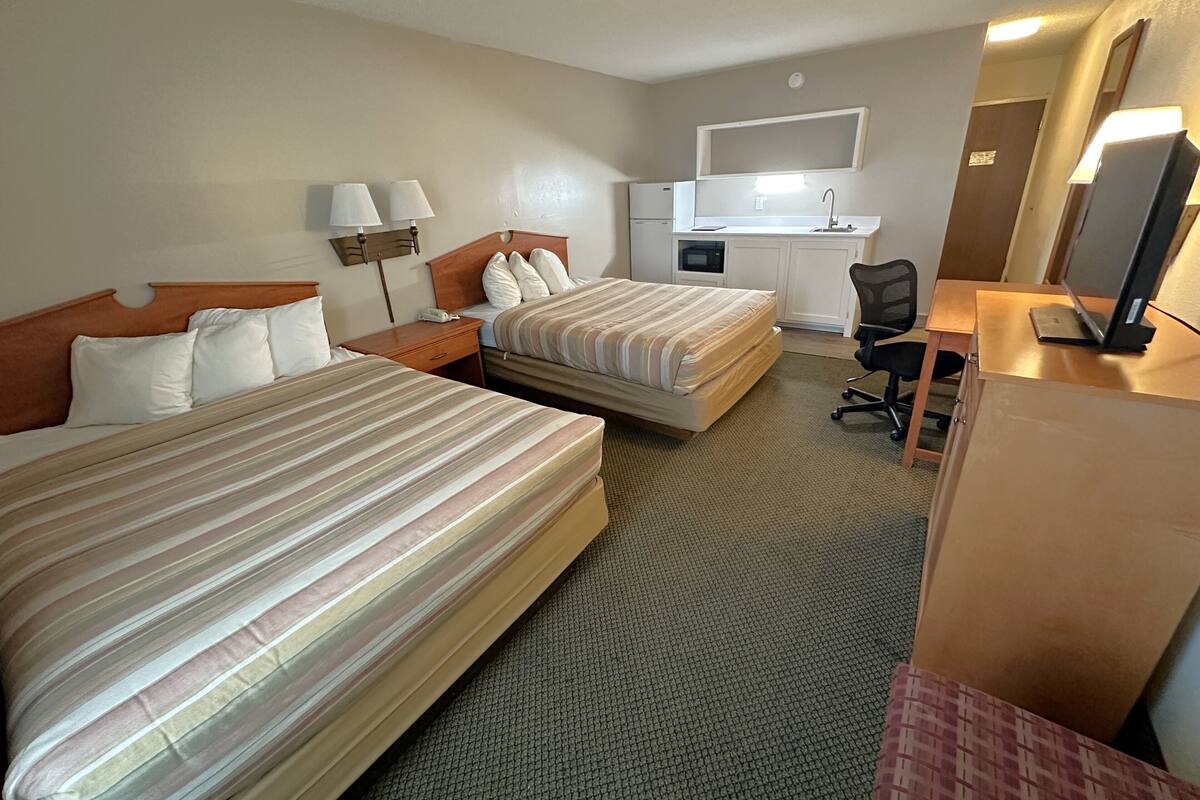 A hotel room with two beds, a desk, a chair, a small kitchenette, and a TV.