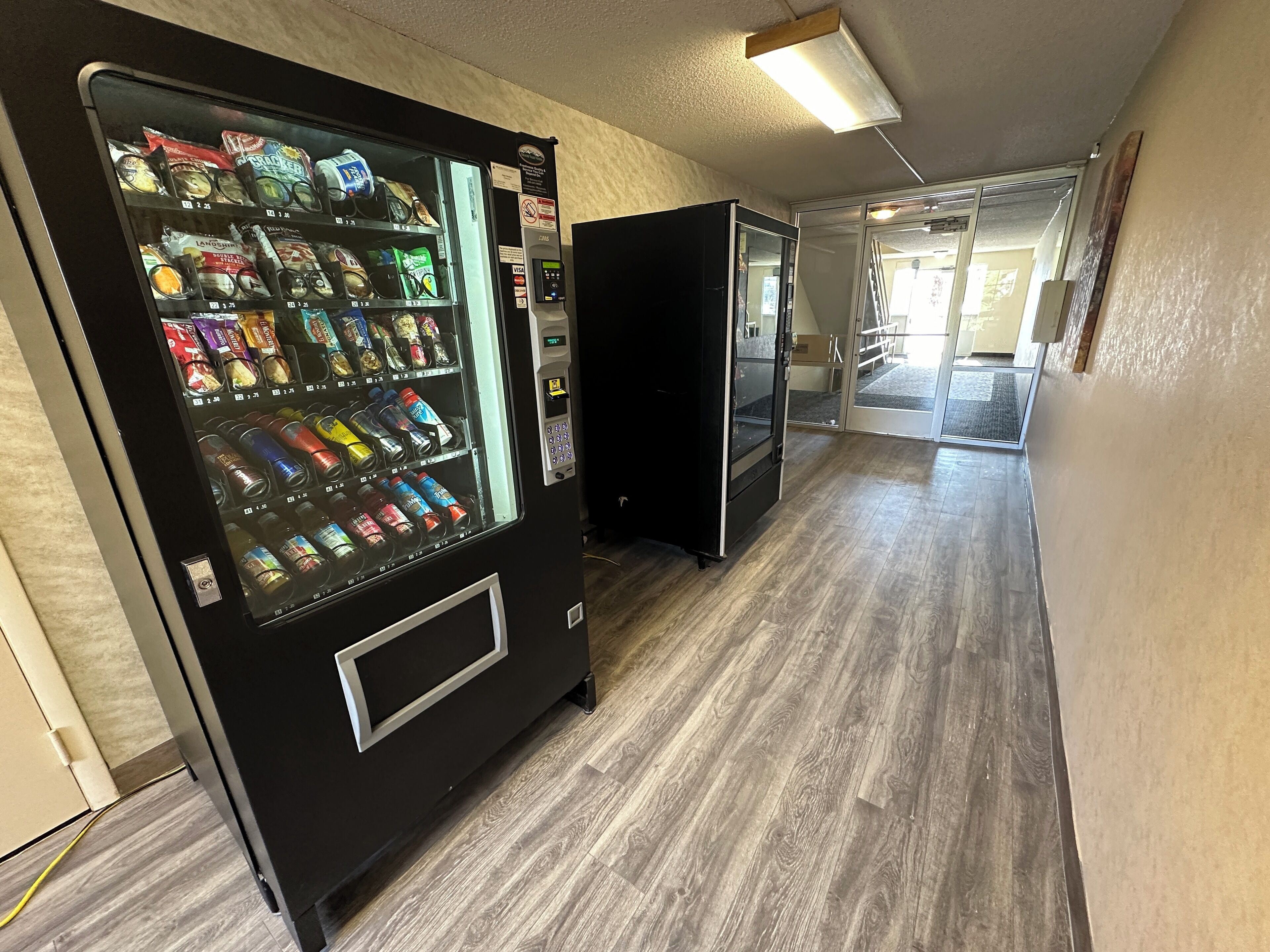 A hallway with a vending machine stocked with snacks and drinks, a black refrigerator, and a wooden floor.