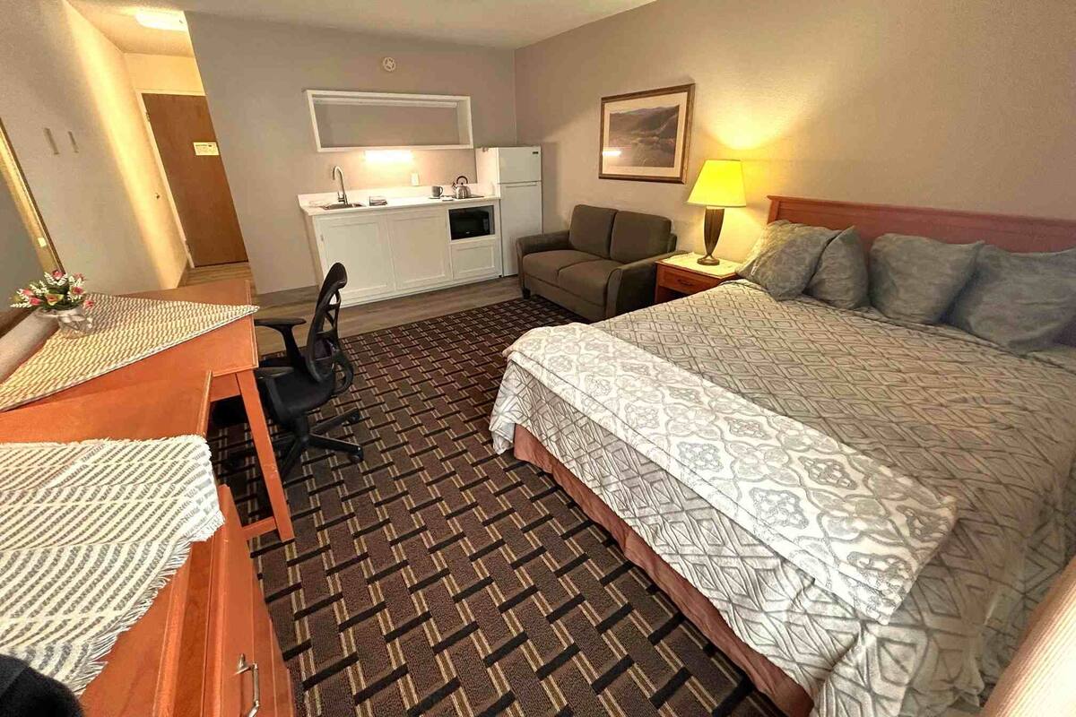 A hotel room with a bed, a desk, a chair, a sofa, a small kitchenette, and a lamp.