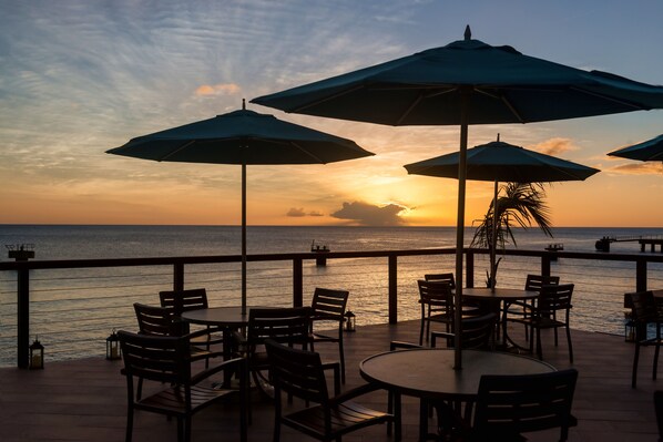 Una terraza con mesas y sillas, sombrillas y vistas al atardecer sobre el océano.