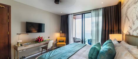 Une chambre d’hôtel moderne dotée d’un grand lit, d’un bureau avec un ordinateur, d’un téléviseur fixé au mur et d’un balcon offrant une vue sur la mer.