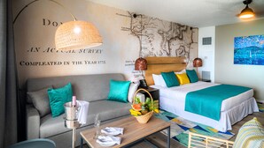 Une chambre d’hôtel avec un mur décoré d’une carte, un lit, un canapé, une table basse sur laquelle se trouve un panier de fruits, et une lampe.