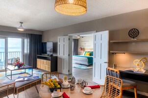 Habitación de hotel con zona de comedor, cama con cubrecama azul y blanco, televisor y vista al océano.