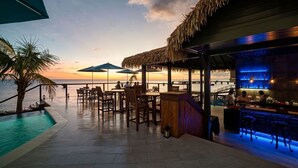 Un bar en bord de mer avec une terrasse, une piscine et une vue sur l’océan au coucher du soleil.