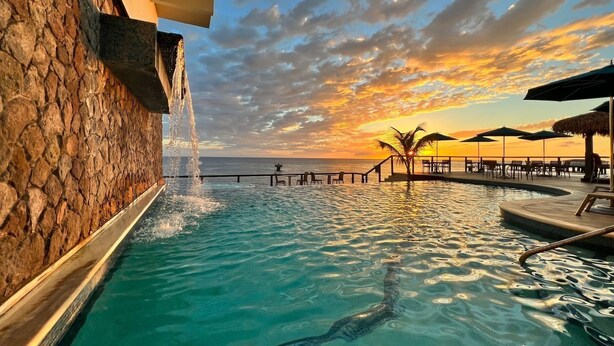 Une piscine agrémentée d’une cascade, offrant une vue sur le coucher de soleil, et un espace de détente avec des parasols.