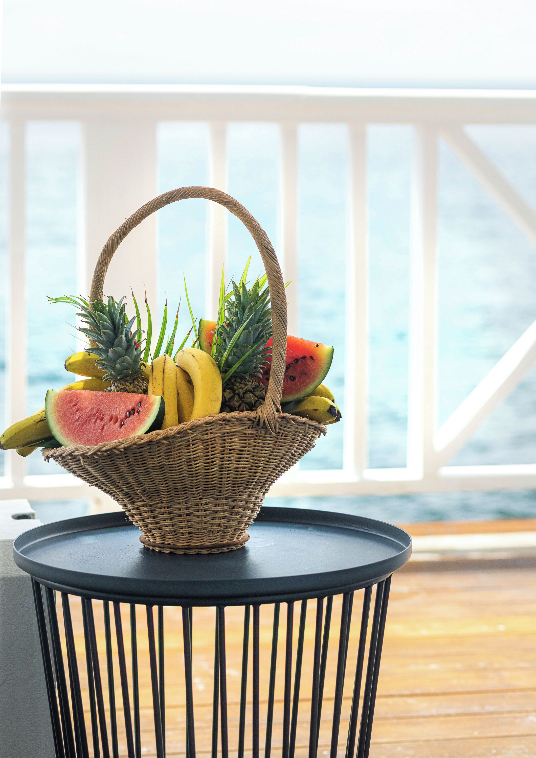 Ein Korb aus Weidengeflecht mit Bananen, Ananas und Wassermelone auf einem runden Tisch mit schwarzem Metallgestell, platziert auf einer Holzdiele mit Geländer und Blick auf den Ozean.
