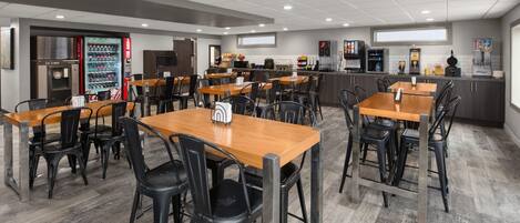 Una cafetería moderna con mesas de madera y sillas negras, equipada con máquinas expendedoras y una estación de café.