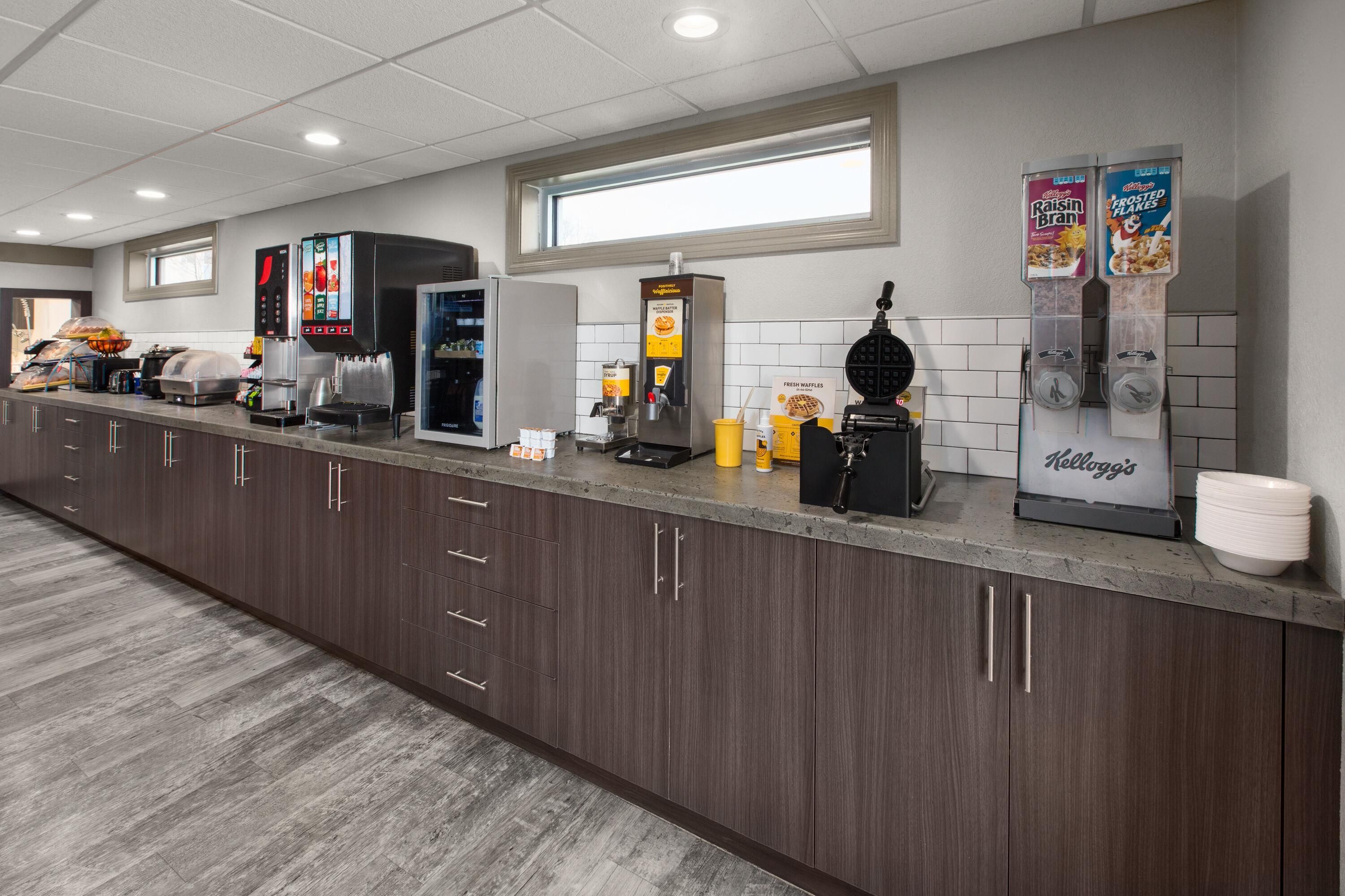 A hotel kitchen with a self-service breakfast bar, including a coffee machine, waffle maker, and various food dispensers.