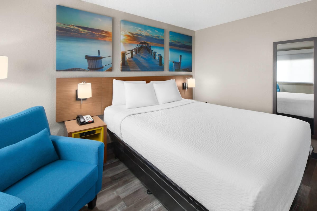 Habitación de hotel con cama, un sillón azul, una mesita de noche con teléfono y decoración en la pared que representa un muelle y un atardecer.