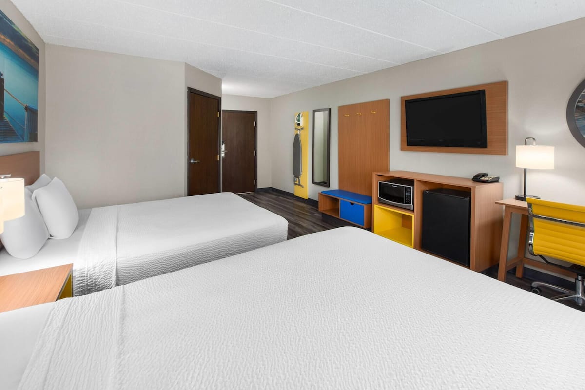 Habitación de hotel con dos camas, un escritorio con lámpara, televisor, espejo y un refrigerador pequeño.