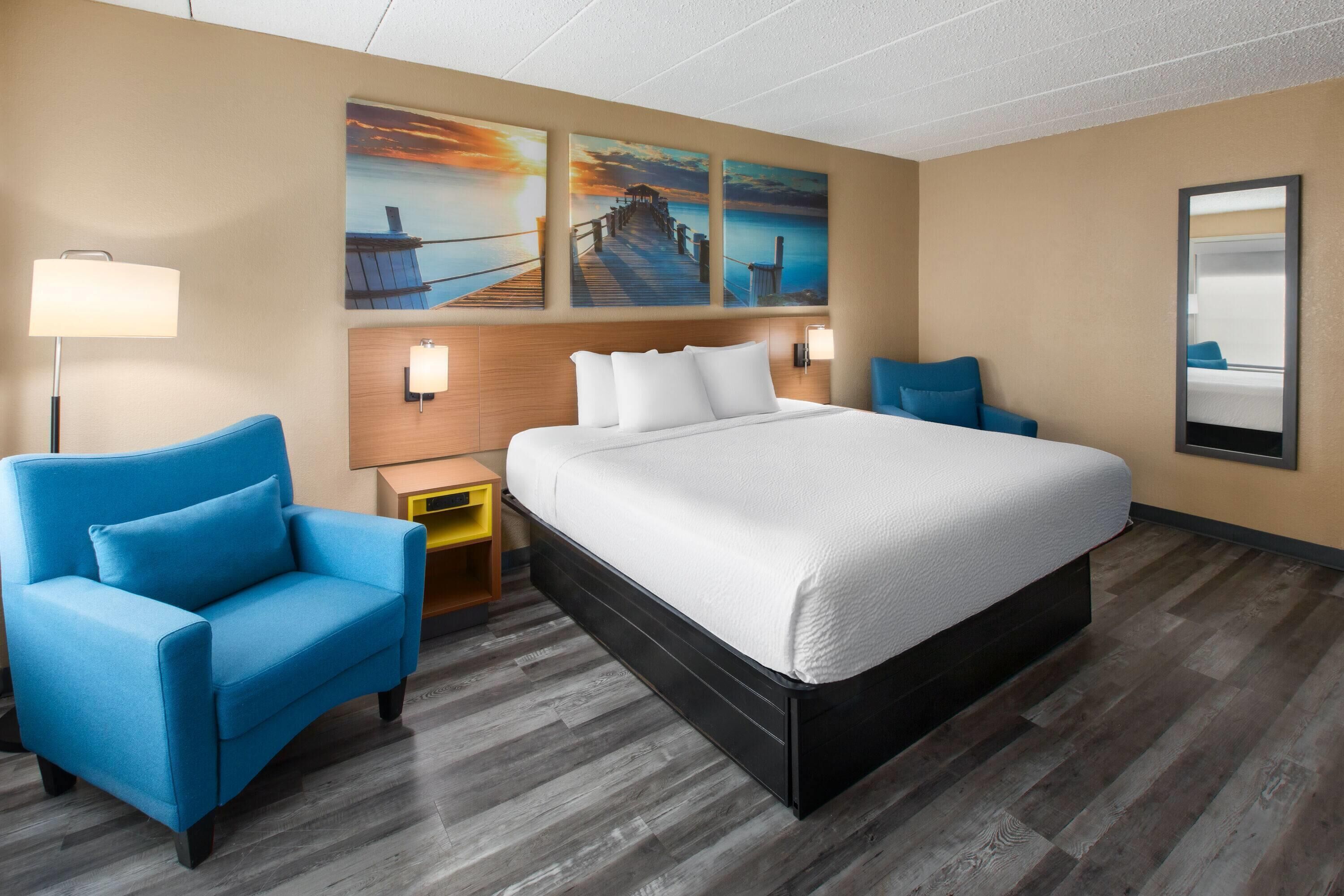 Hotelkamer met een groot bed, twee blauwe fauteuils, een houten hoofdbord, een nachtkastje, een lamp en drie aan de muur bevestigde schilderijen.