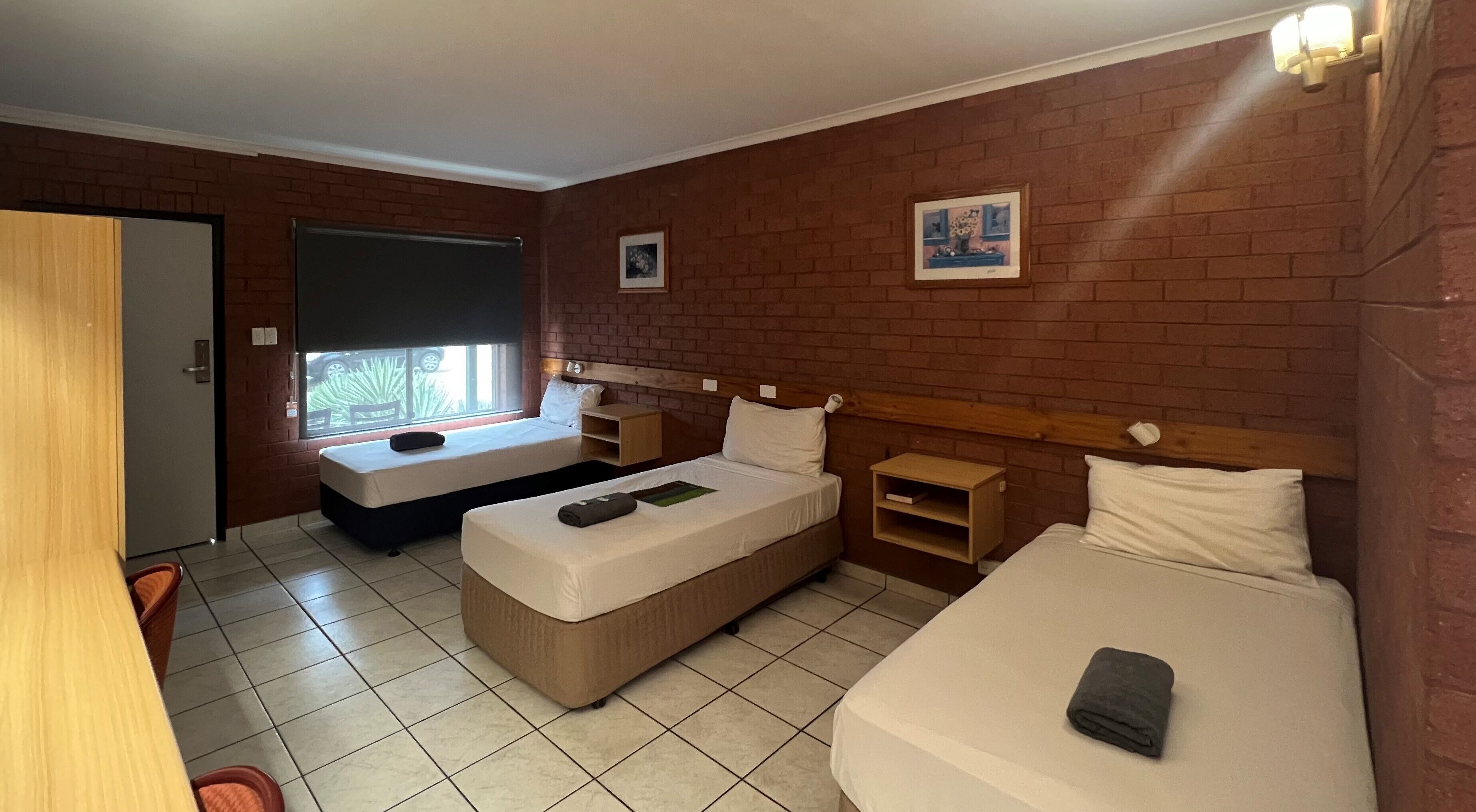 A hotel room with two single beds, a sofa bed, a small wooden nightstand, a window with a view of a car, and a brick wall.