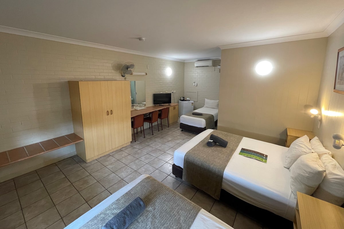 Una habitación de hotel compacta con dos camas individuales, un escritorio pequeño con una silla, televisor y un área de kitchenette.
