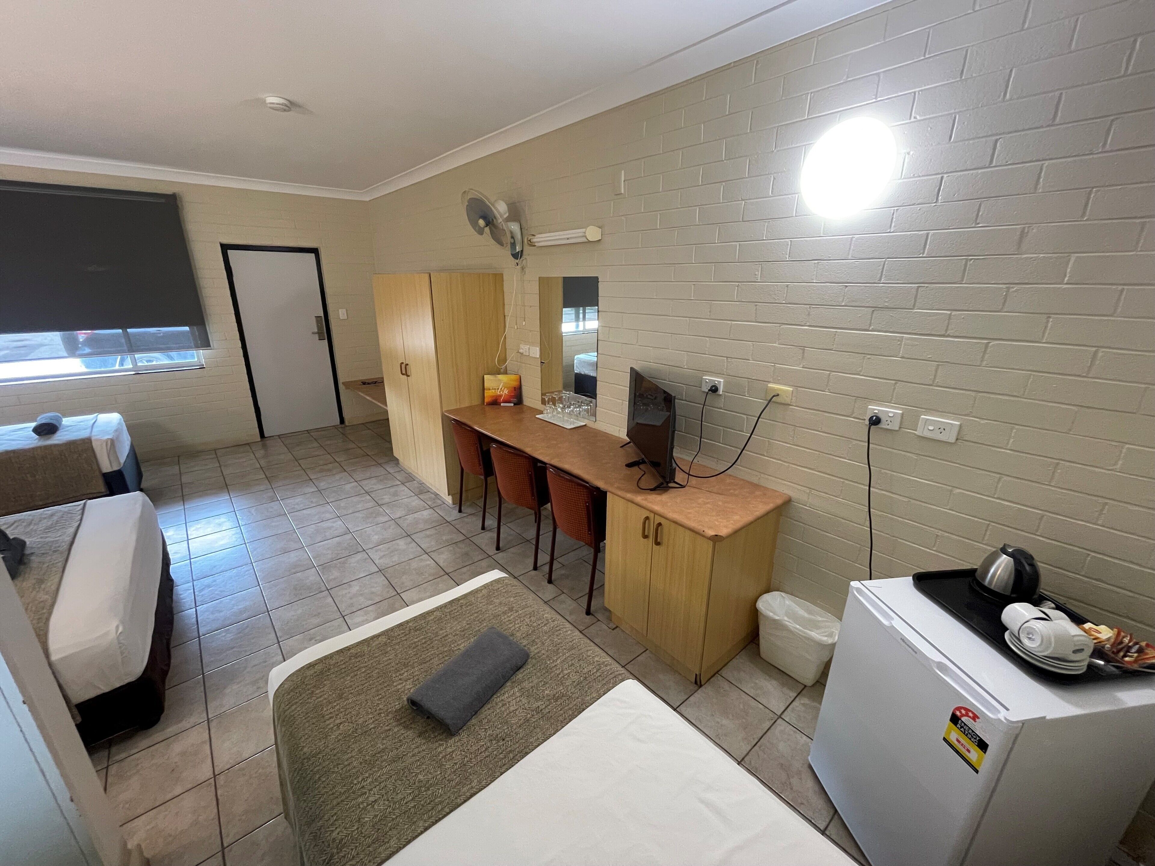 A compact hotel room with a bed, a desk with a computer, a small refrigerator, and a window with blinds.