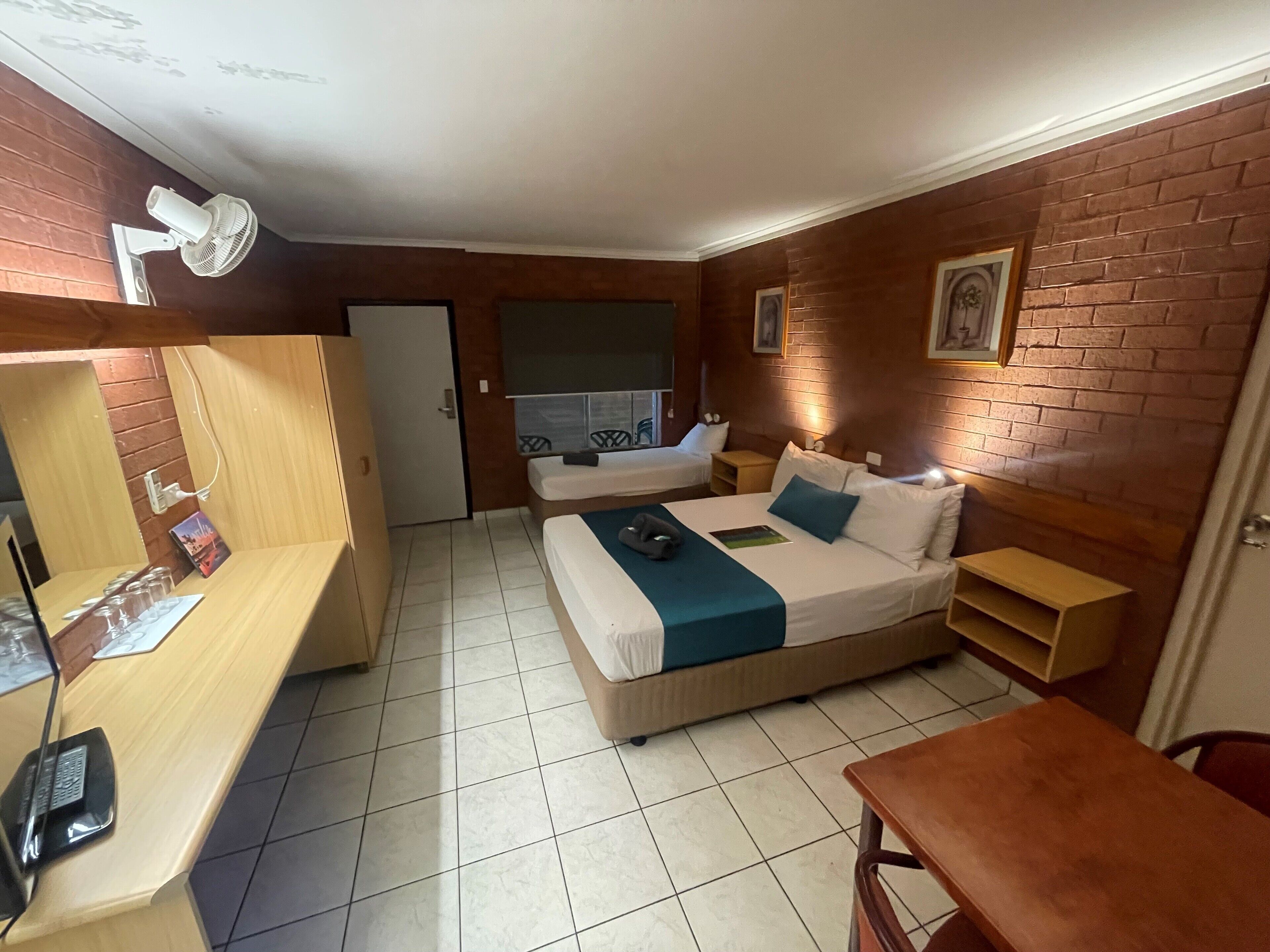 A hotel room with a bed, bedside tables, a desk with a chair, a mirror, and a fan.