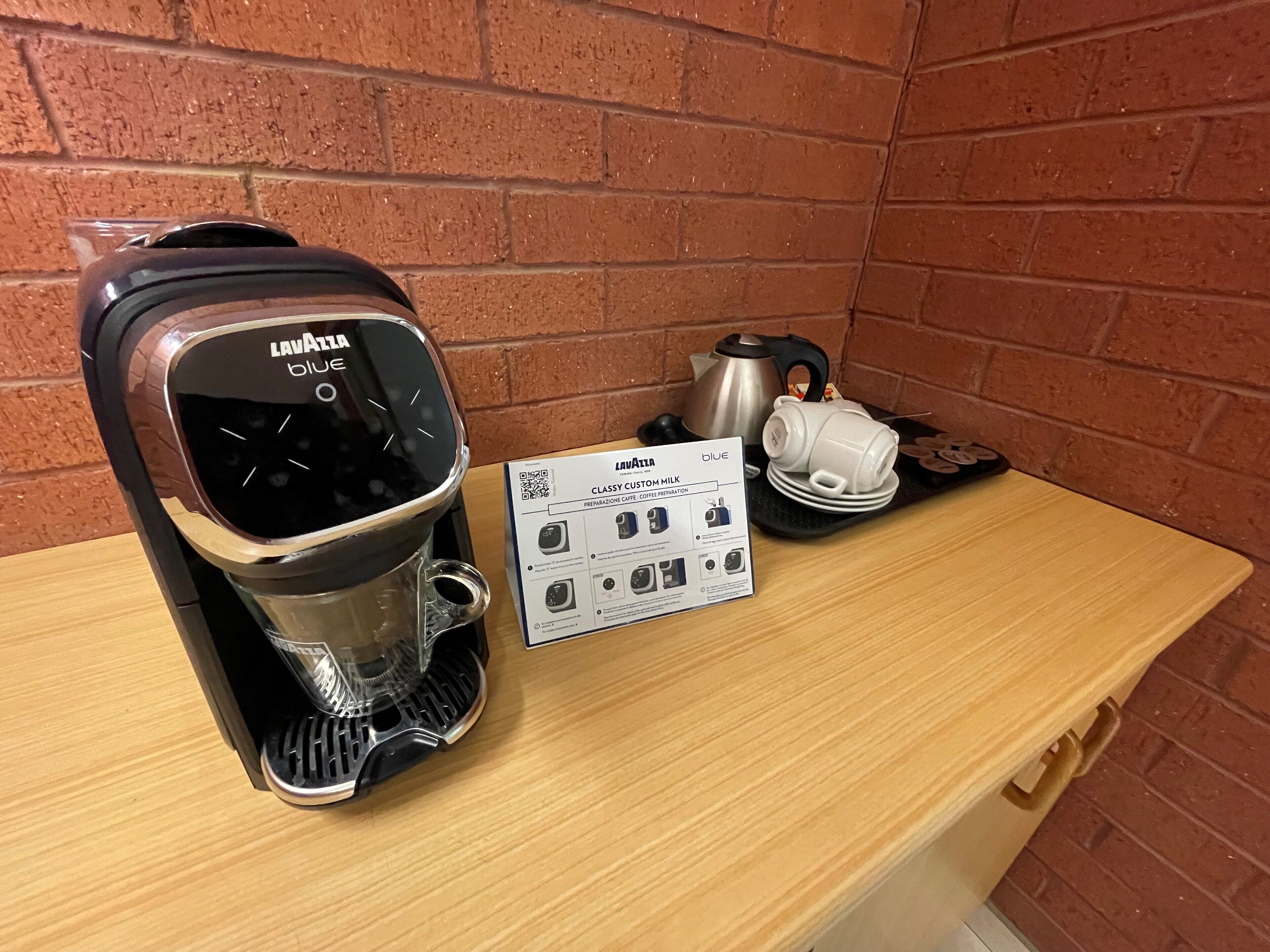 Una cafetera Lavazza con una moka y una jarra de café sobre una mesa de madera frente a una pared de ladrillo.