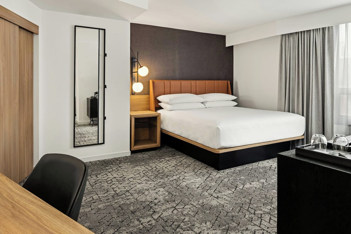 A hotel room with a large bed, a desk, a chair, a mirror, a nightstand, and a lamp.