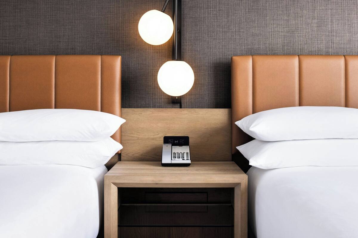 A hotel room with two beds, a wooden nightstand with a phone, and two wall-mounted lights.