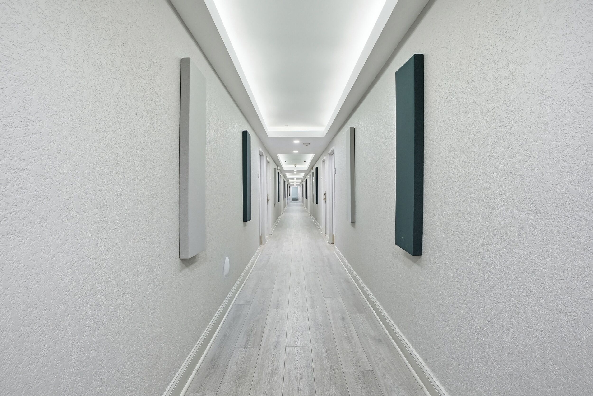 A long, narrow hallway with white walls and a light wooden floor, leading to a bright, well-lit end.