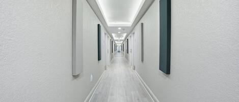 A long, narrow hallway with white walls and a light wooden floor, leading to a bright, well-lit end.
