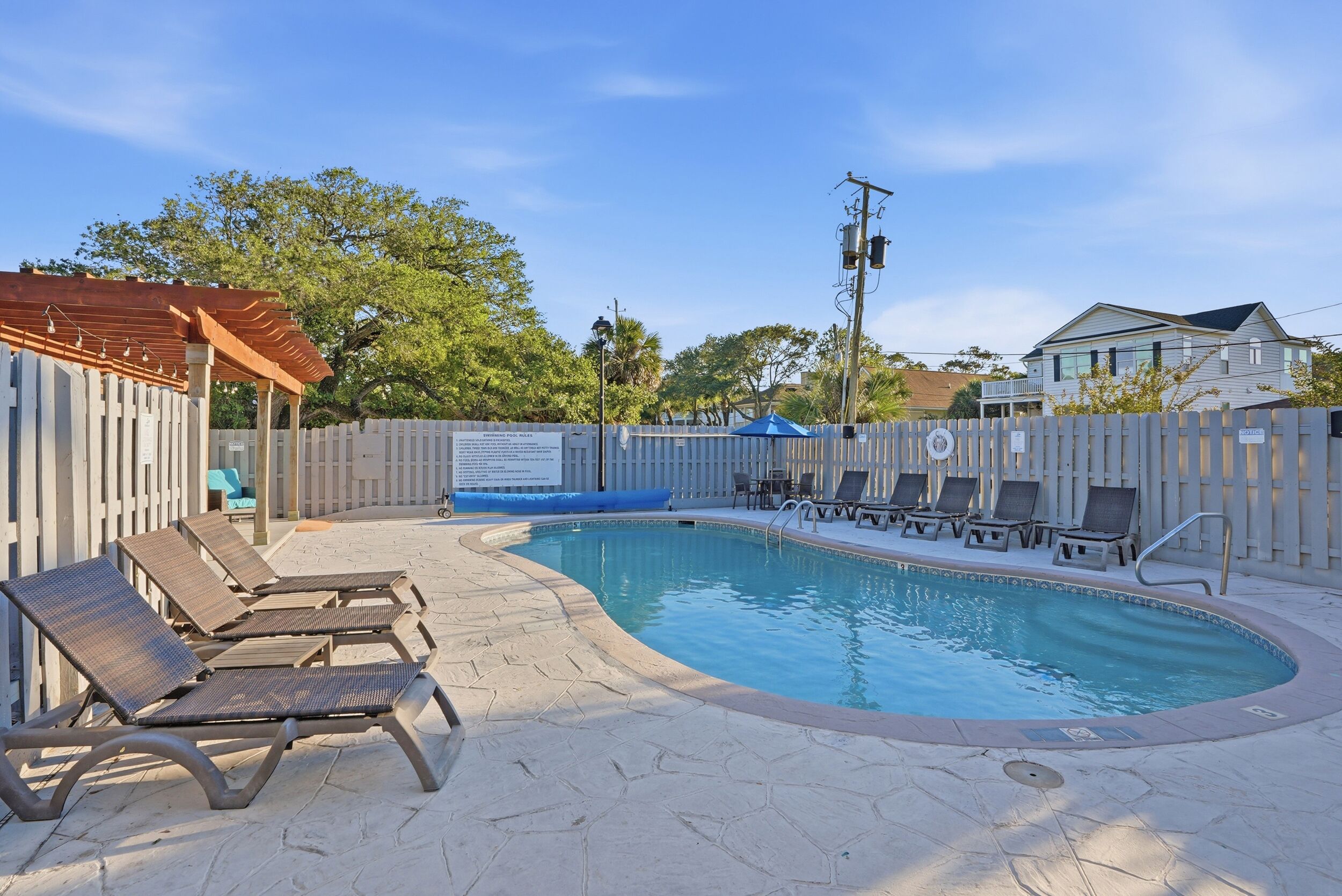 A swimming pool with lounge chairs, a pergola, and a fenced area with a sign.