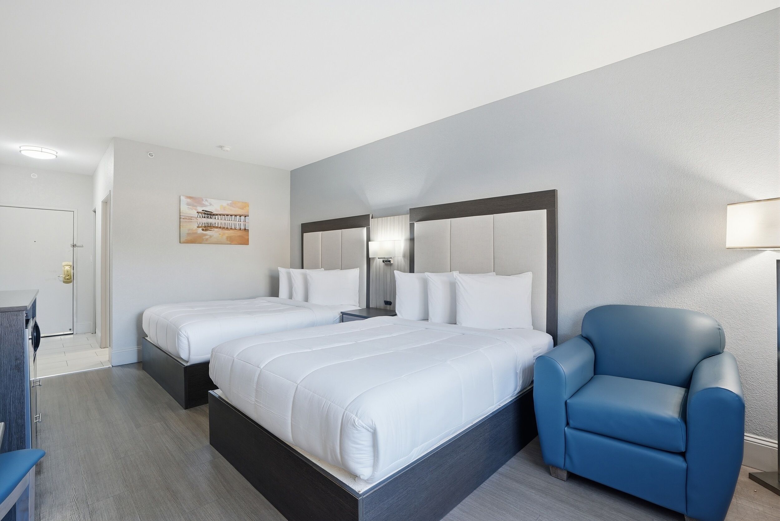 Una habitación de hotel con dos camas, un sillón azul, un escritorio y un cuadro en la pared.