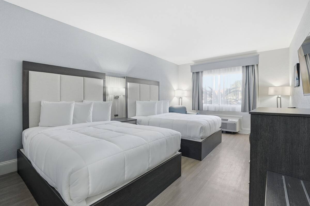 A hotel room with two beds, a desk, a chair, a window with curtains, and a view of the city.
