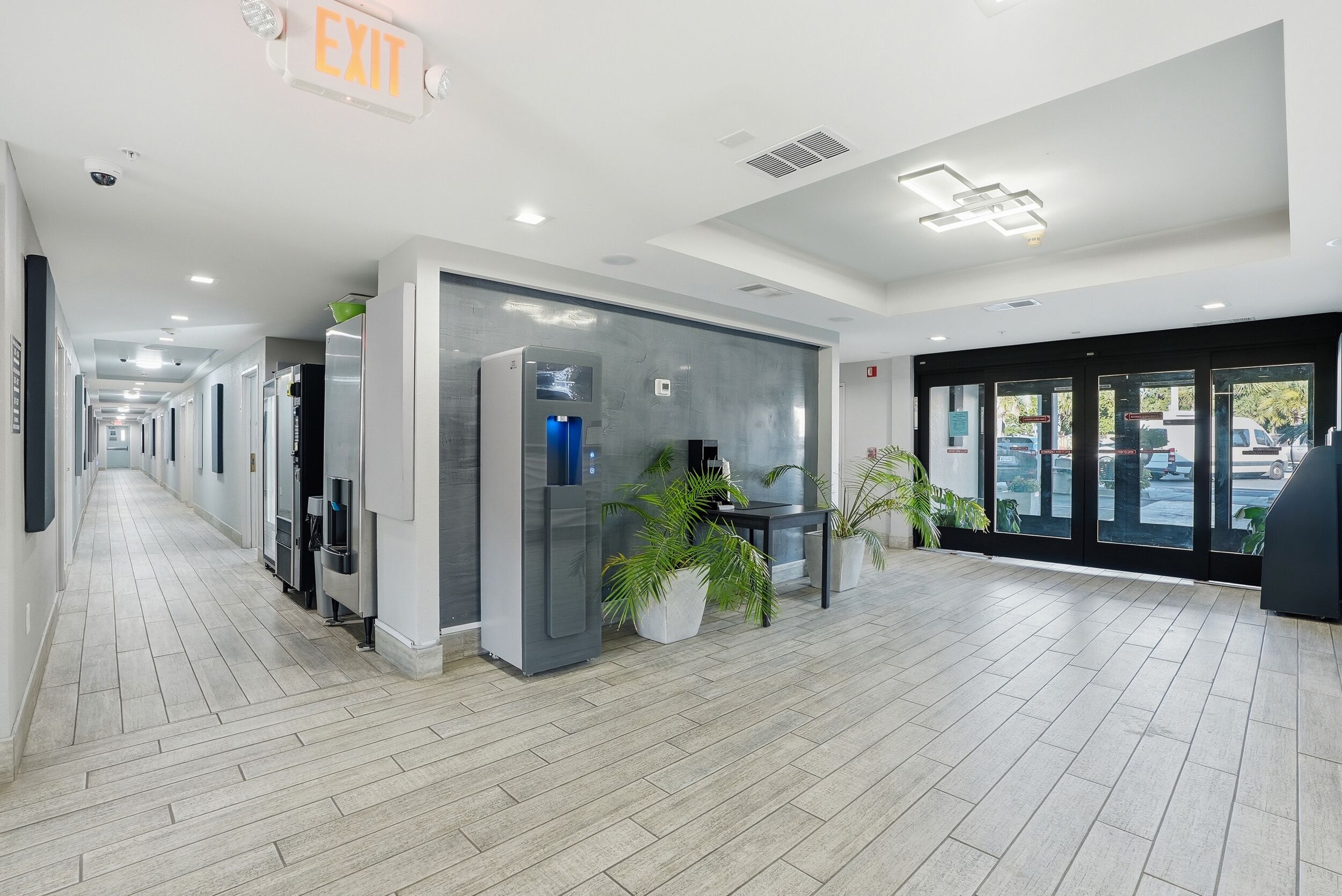 A hotel lobby with a long corridor, an exit sign, a water dispenser, and potted plants.