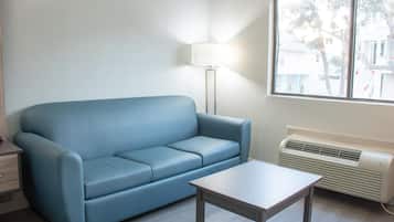 Una habitación con un sofá azul, una mesita, una lámpara de pie y un ventanal con vistas a edificios y árboles.