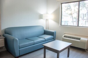 Una habitación con un sofá azul, una mesita, una lámpara de pie y un ventanal con vistas a edificios y árboles.