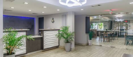 A modern hotel lobby with a reception desk, potted plants, and a dining area with tables and chairs.