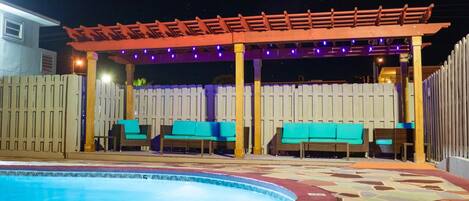 A pool area with a wooden pergola and seating under string lights.