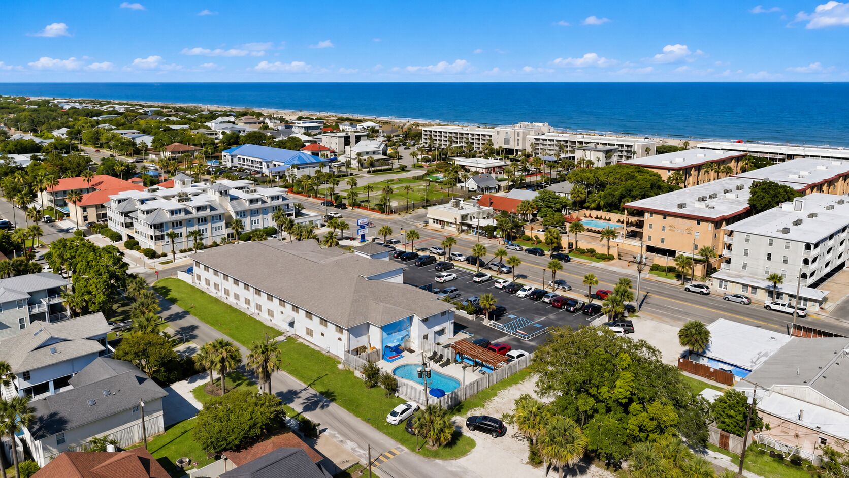 An aerial view of a coastal town with a hotel featuring a swimming pool, multiple buildings, and a clear view of the ocean.